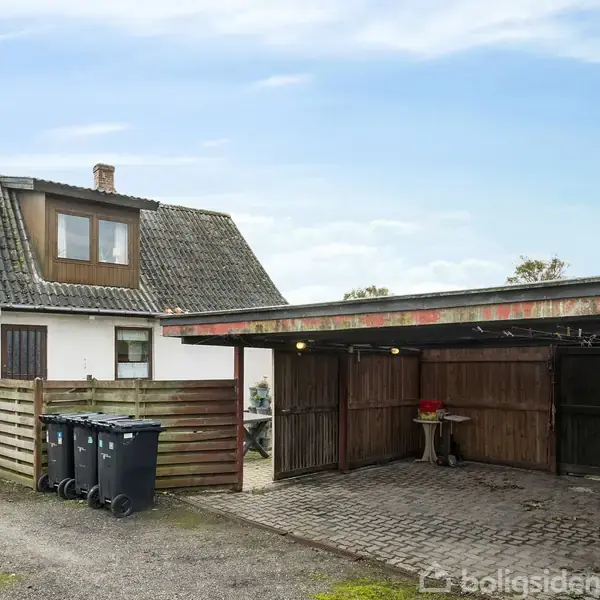 Hus med skråtag og hems står stille ved siden af en carport. Tre skraldespande står foran et træhegn. Grusvej løber langs ejendommen med busk i højre side.