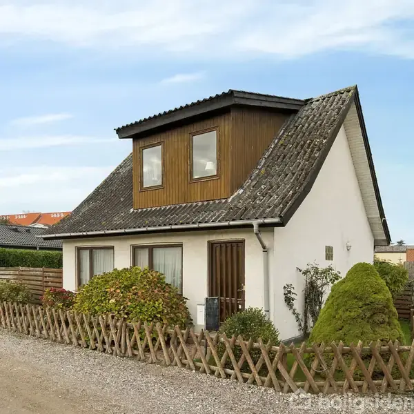 Et hvidt hus med en brun kvist på taget står i en grusvej omgivet af en lav trælignende hegn. Buske og en sti er foran huset.