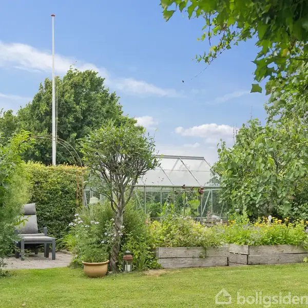 Grønt område med en pavillon, omgivet af frodige planter og buske. Et flagstang står i baggrunden, og en hvid drivhus er synlig blandt træerne.