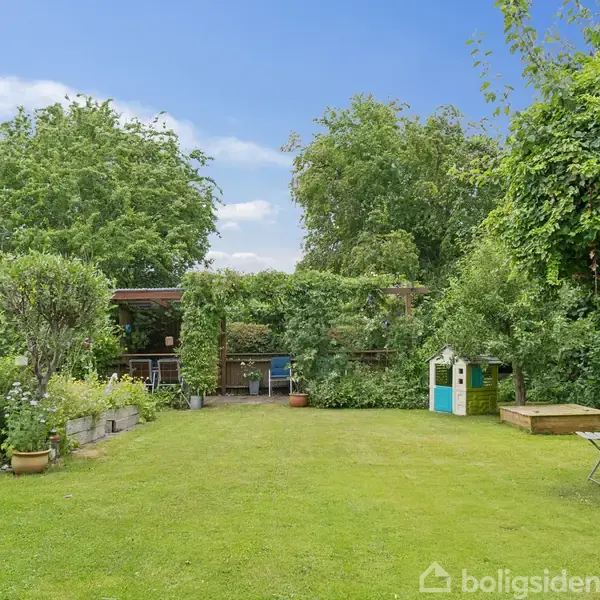 En grøn plæne er omkranset af frodige buske og træer. Til højre står et lille legehus, og til venstre ses blomsterkrukker og en terrasse med havemøbler under en pergola.