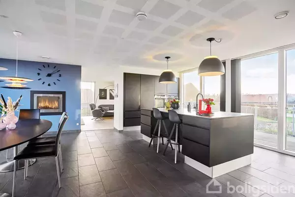 Køkkenø med to barstole, oplyst af hængelamper, står i et moderne køkken med store vinduer. Til venstre ses en spisestue med pejs, blomsterdekorationer og vægklokke.