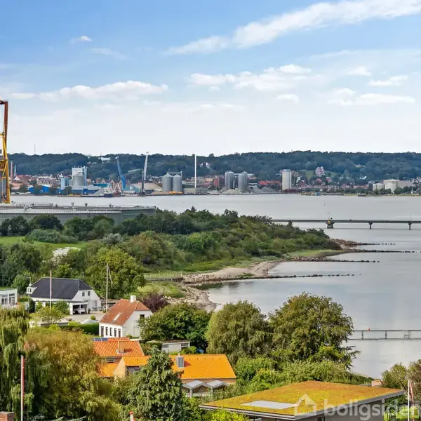 Huse med rødt tage ligger langs kysten i en frodig landsby. I baggrunden ses en stor kran og en havnefront over en bred flod med en bro.