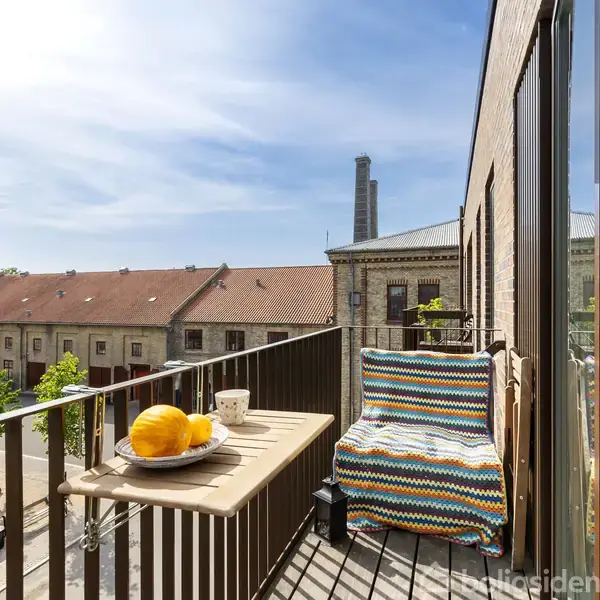 En balkon med et lille bord og en farverig stol. Bordet har en tallerken med appelsiner og en kop. Omgivet af bygninger og en blå himmel.