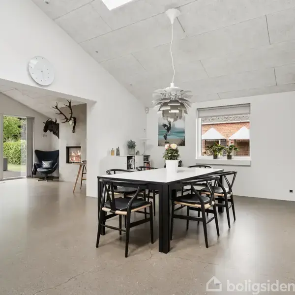 Spisestue med sort bord og seks stole, dekoreret med planter på bordet. I baggrunden ses en pejs, vægdekorationer af dyrehoveder og en stor glasdør mod en terrasse og have.