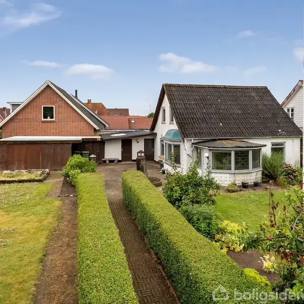 Hus med mørkt tag står ved siden af en grussti og velplejet hæk. I baggrunden ses en garage samt andre huse i et villakvarter med grønne områder.