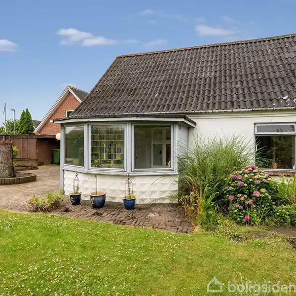 Et hvidt hus med skråt tag står foran en frodig have med græsplæne og farverige blomster. En brolagt terrasse ses ved siden af huset med blomsterkrukker foran.