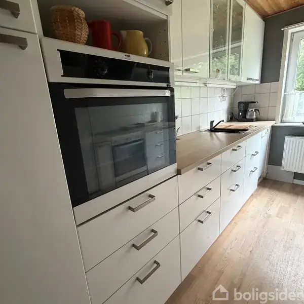 En moderne komfur stående i et køkken, omgivet af hvide skabe og en træbordplade. Kaffen er lavet med en kaffemaskine, og en vask ses nær et vindue med udsigt.