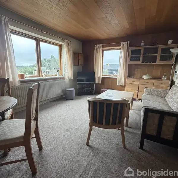 Stue med træmøbler, sofa og spisebord placeret ved vinduer, som viser udsigt til det grønne. Væggene er pyntet med hylder, og gardiner hænger ved vinduerne.