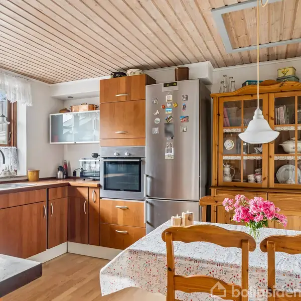 Køkkenbord med blomstret dug, farverige blomster i vase øverst, omgivet af træstole. Køleskab med magneter, bagved. Skab, ovn, og vask ved vinduet i lyse træpanelkøkken.