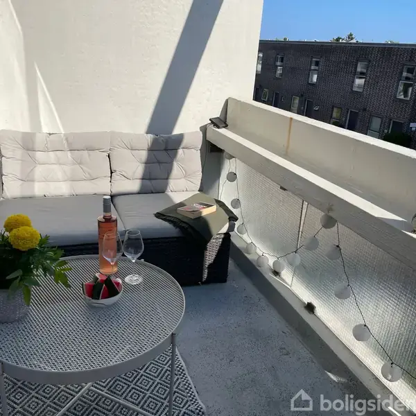 En hyggelig altan med en grå hjørnesofa, et lille rundt bord med blomster, en flaske rosévin og glas. En ranke af lyskæder hænger på gelænderet med udsigt til en bygning.