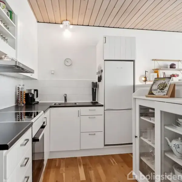 Hvidt køkken med sorte bordplader og moderne apparater, som et køleskab og ovn. En hylde med indrammet kunst og dekorationer. Trægulv og loftslampe supplerer det lyse interiør.