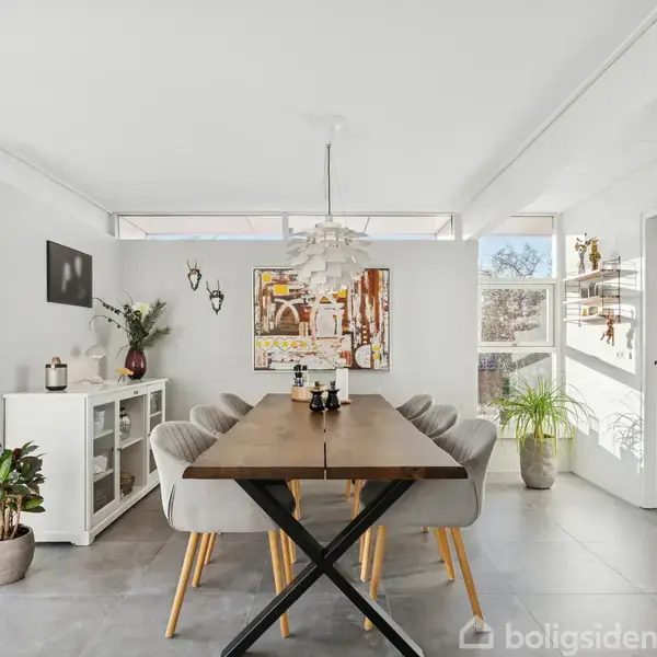 Et spisebord er omgivet af seks stole i en lys spisestue med vinduer til venstre og højre. Vægkunst og planter dekorerer rummet. En dør åbner til haven.