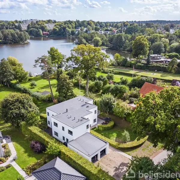 Hvid moderne hus ligger ved siden af en sø omgivet af frodig grøn beplantning og andre huse. I baggrunden ses en rolig skovklædt landskab, der strækker sig ind i horisonten.