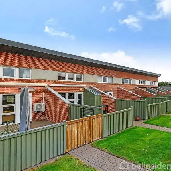 Rækkehuse i røde mursten står på række med grønne små forhaver foran, adskilt af lave hegn. En flisebelagt sti løber langs husene under en blå himmel.