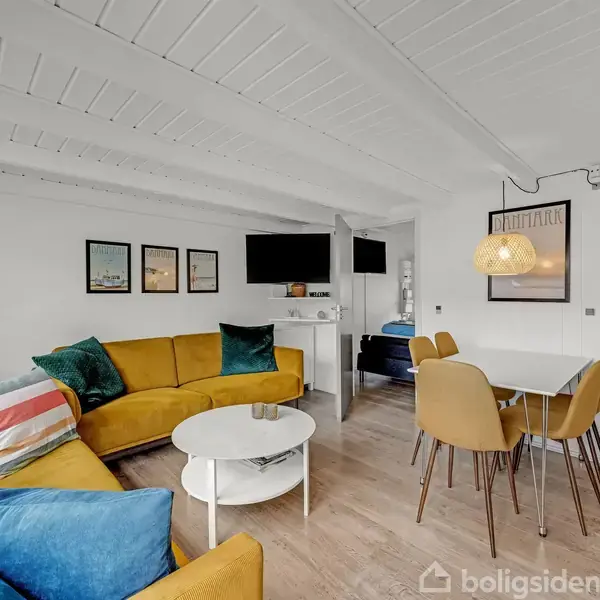 En stue med en gul hjørnesofa fyldt med farverige puder står ved et rundt sofabord. Ved siden af er et spisebord med stole. Væggene er dekoreret med billeder.