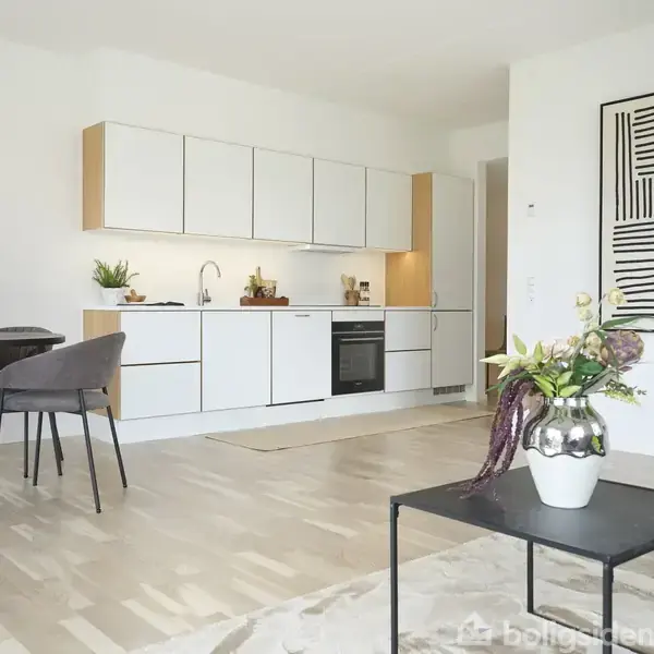 Køkken med hvide skabe og indbygget ovn, ledsaget af et spisebord med stole. En plante på et bord tilføjer dekoration. Minimalistisk kunst pryder den hvide væg.