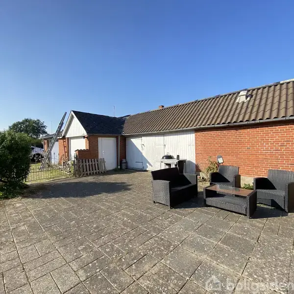 Havemøbler er arrangeret omkring et bord på en flisebelagt terrasse foran en rød murstensbygning. Solskin lyser området op, og en hæk afgrænser terrassen til venstre.