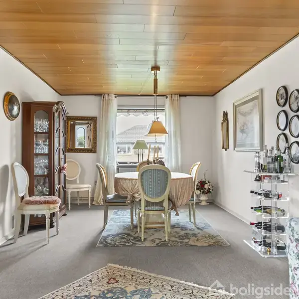 Spisebord dækket med en dug, omgivet af stole i en stue med lyse vægge. En lysestage og malerier hænger på væggen, tæpper dækker gulvet, og et vitrineskab står i hjørnet.