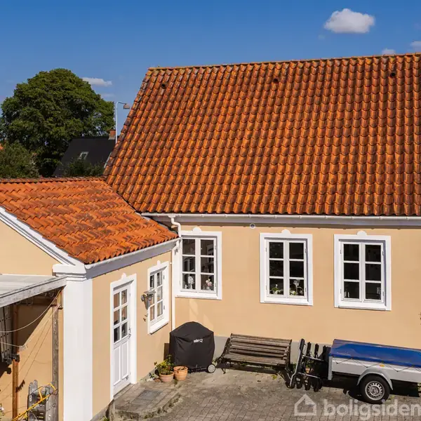 Et gult hus med rødt tegltag står stille i en solrig gårdsplads. Ved siden af døren står en grill og en trailer. Himlen er klar og blå.