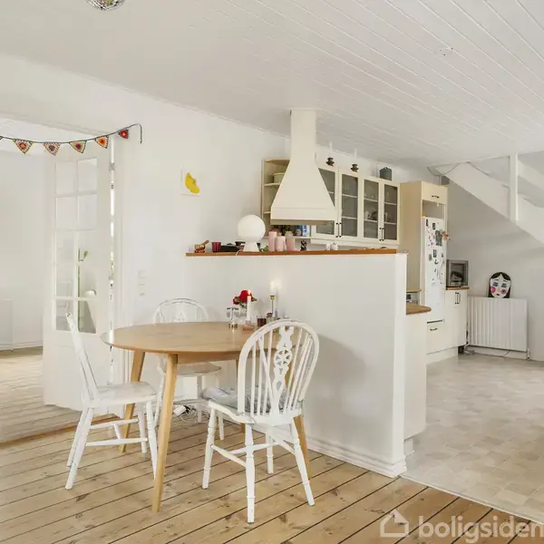 Et gult hus med rødt tegltag står stille i en have omgivet af hække og grussti. Flere huse og et glimt af vand ses i baggrunden.