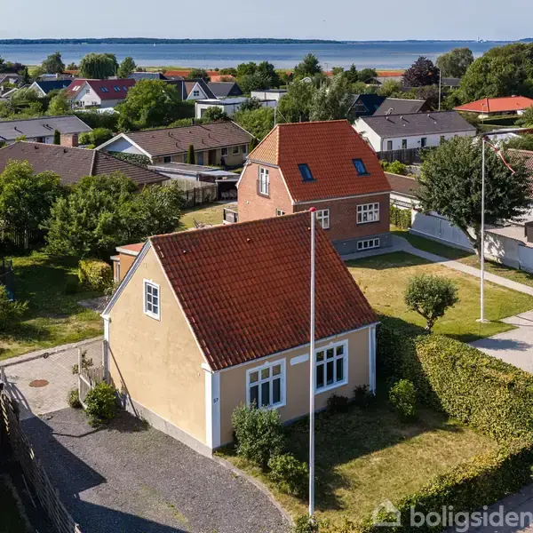 Et hus med rødt tag og to vinduer står i en forstadshave med hække og træer, mens flere huse og en flod ses i baggrunden.