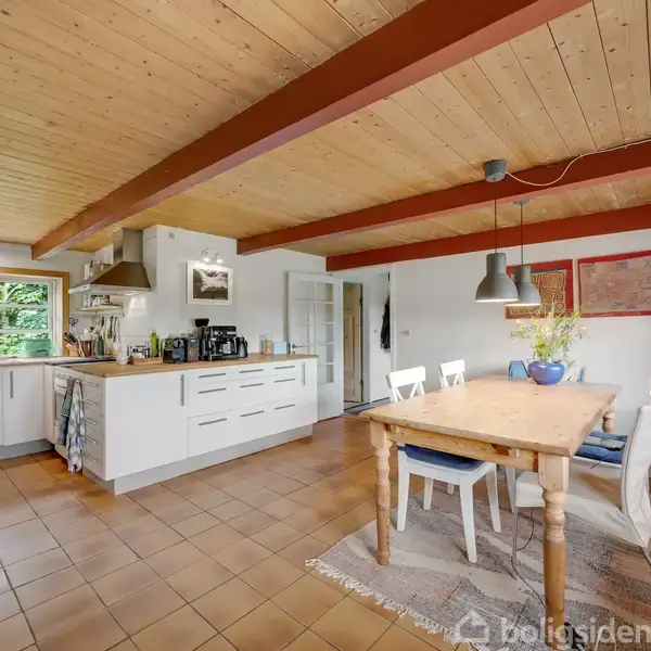 Køkken med træloft og fliser; et bord med stole står på et tæppe, og der hænger lamper over bordet. Store vinduer giver lys til rummet, som er lyst og åbent.