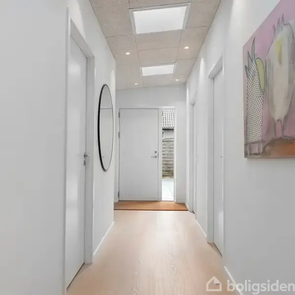 En lys gang med trægulv, hvor dørene står lukket på begge sider. Et rundt spejl hænger på venstre væg, mens et abstrakt maleri er til højre.