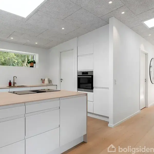 Et moderne køkken med hvide elementer og en lys træbordplade. Der er indbygget ovn i væggen, planter på bordet, et vindue og en gang med rundt spejl i baggrunden.