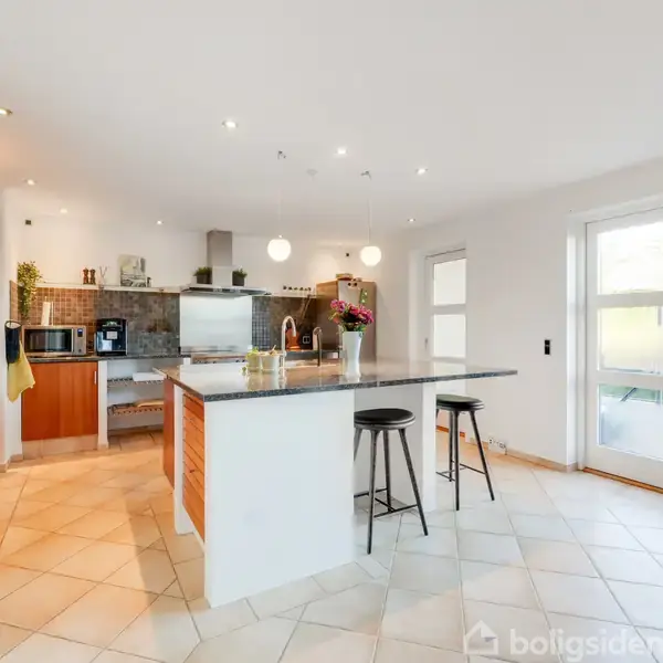 Et moderne køkken med en stor ø i midten, omgivet af barstole, står oplyst af indbyggede lys. Baggrunden har køkkenapparater og åbne hylder med planter.