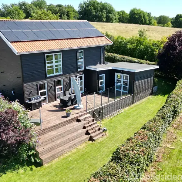 Et sortmalet hus med solpaneler på taget står stille i et grønt landskab. En træterrasse med havemøbler breder sig foran huset, omgivet af buske og græsplæner.