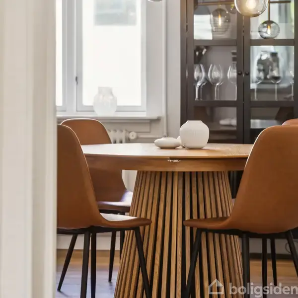 Et rundt træbord omringet af brune stole står i en spisestue med vindue. Bordet er pyntet med en hvid vase, og et vitrineskab med glas er i baggrunden.