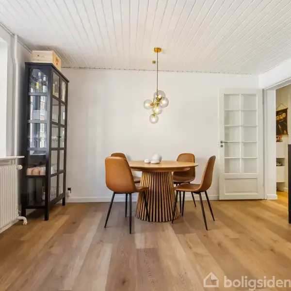 Rundt spisebord med fire brune stole står i et lyst rum med trægulv. En moderne lampe hænger over bordet. Der er en glasvitrine til venstre og en dør i baggrunden.