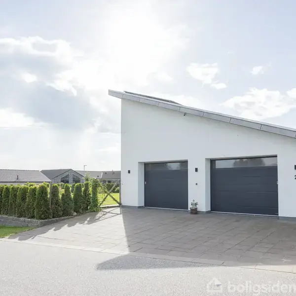 En moderne hvid bygning med to store garagedøre står stationær i et boligområde. Hegn og buske omkranser ejendommen. Nummeret "2" er synligt ved den højre garage.