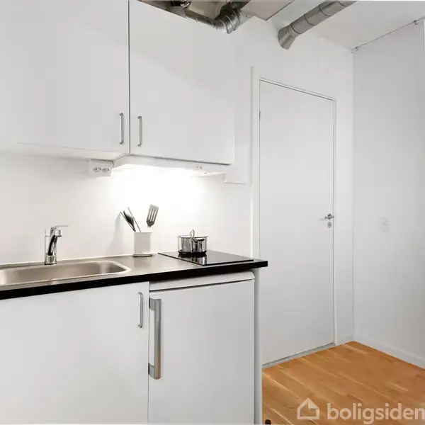 Hvidt køkken med vask, armatur og mørk bordplade. Ovenpå skabet hænger knive i en holder. Rummet har lyst trægulv og døre til tilstødende rum.