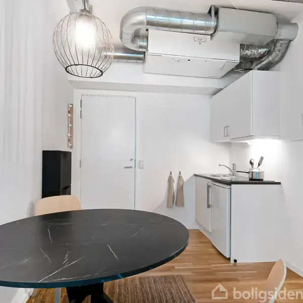 Rundt sort spisebord med stol står i et lille køkken. Køkkenet har en vask, komfur, hvidt skab og hvid væg. Ovenfor ses udsugningskanaler. En lampe hænger fra loftet.