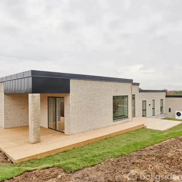 Et moderne, enkelt-etagers hus står stille på en græsplæne. Huset har en lys murstensfacade og store vinduer. Landskabet omkring er åbent med marker og nogle træer i baggrunden.