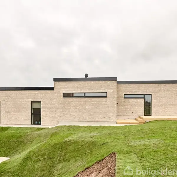 Minimalistisk moderne hus med fladt tag og lyse mursten, står i et grønt, kuperet landskab. Store vinduer giver lysindfald. Skyet himmel i baggrunden indikerer overskyet vejr.