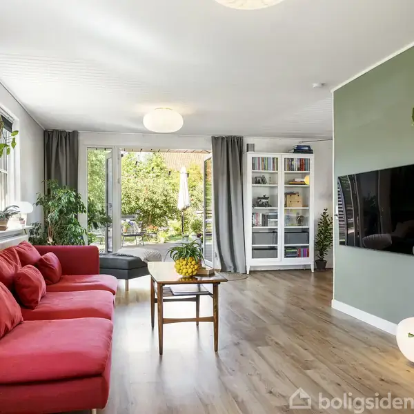 Rød sofa står i stue med grønne planter og træbord. Væggen har tv og bogreol. Store vinduer med gardiner åbner ud til have med lysindfald.