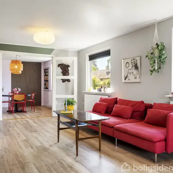 Rød sofa står i en lys stue med trægulv, flankeret af planter. Gennem en åbning ses en spisestue med røde stole. Væggen har kunst og vinduer med udsigt til træer.
