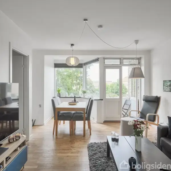 Et moderne stueområde med en stor fladskærms-tv til venstre, sofagruppe med sofabord til højre, og spisebord foran et vindue med udsigt til grønne omgivelser.