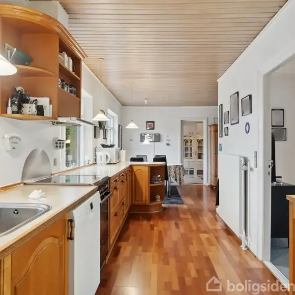 Køkken med træskabe og en lang, smal bordplade, udstyret med vask og ovn. Moderne apparater er synlige. Store vinduer giver naturligt lys, og der er en dør til en tilstødende stue.