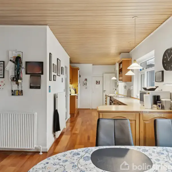 Køkken-alrum med trægulv og lyst loft. Rundt spisebord med mønstret dug og sorte stole. Vægge dekoreret med billeder. Køkkenområdet har hvide skabe, pendellamper og åbner mod en gang.