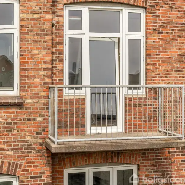 En murstensfacade med en lille balkon prydet af hvide rækværk. Bag balkonen er der store vinduer, der åbner op til en lys lejlighed.