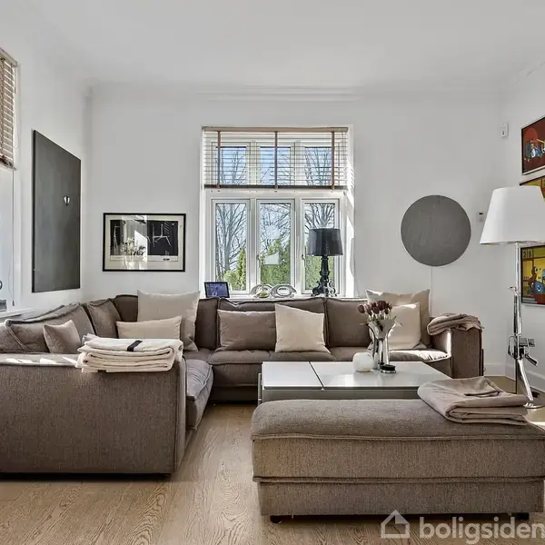En stor, brun sofa står i hjørnet af en skandinavisk stue, omgivet af kunst på væggene og vinduer med lyse gardiner. Indretningen er lys og moderne.