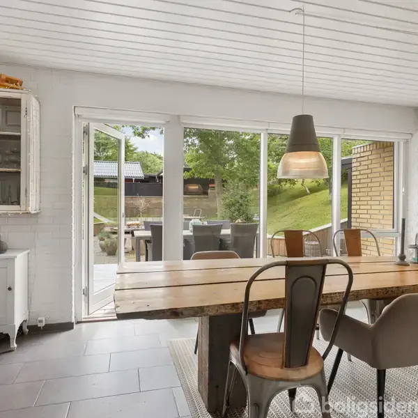 Et træbord med stole står i et spisekøkken. Store vinduer fører ud til en terrasse med grønne omgivelser. To moderne lamper hænger over bordet, og en hylde står i hjørnet.