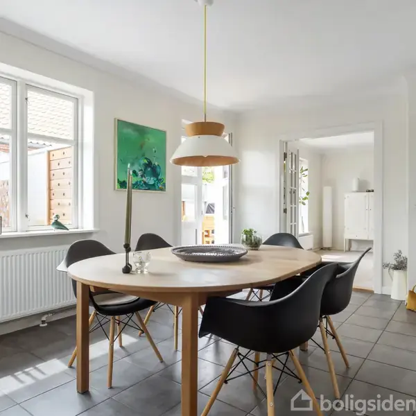 Rundt spisebord med sorte stole omringet af lys grå flisegulv. I baggrunden ses store vinduer med udsigt til en have. En moderne lampe hænger over bordet.
