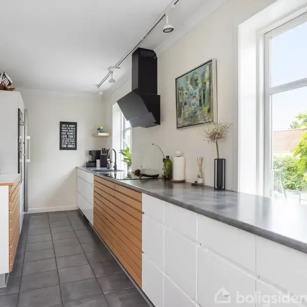Et moderne køkken har hvide og træskuffer, en grå bordplade og adskillige køkkenapparater. Store vinduer slipper lys ind, mens planter dekorerer området. På væggen hænger et billede og en plakat.