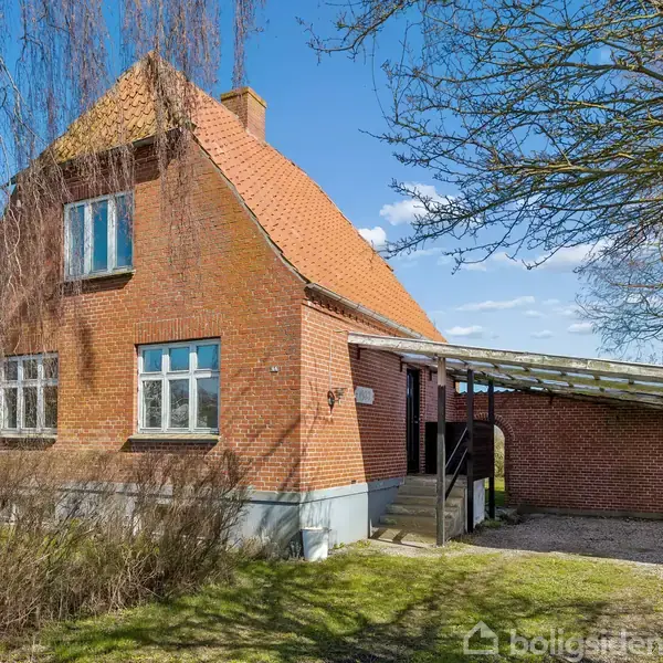 Rødt murstenshus står stille i en have med vinterklædte træer. Huset har et skråt tag og indbygget carport. Skyfri, blå himmel danner baggrund.