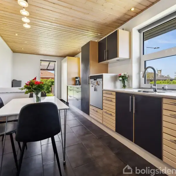 Et moderne køkken har en række sorte og træfarvede skabe, en spiseplads med fire stole og et bord. Et stort vindue giver udsigt til en grøn have.