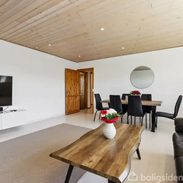 Fladskærms-TV på hvid væg, slukket, i moderne stue med trægulv. Rundt spisebord med sorte stole og spejl. Sofa i forgrunden, træloft overalt.